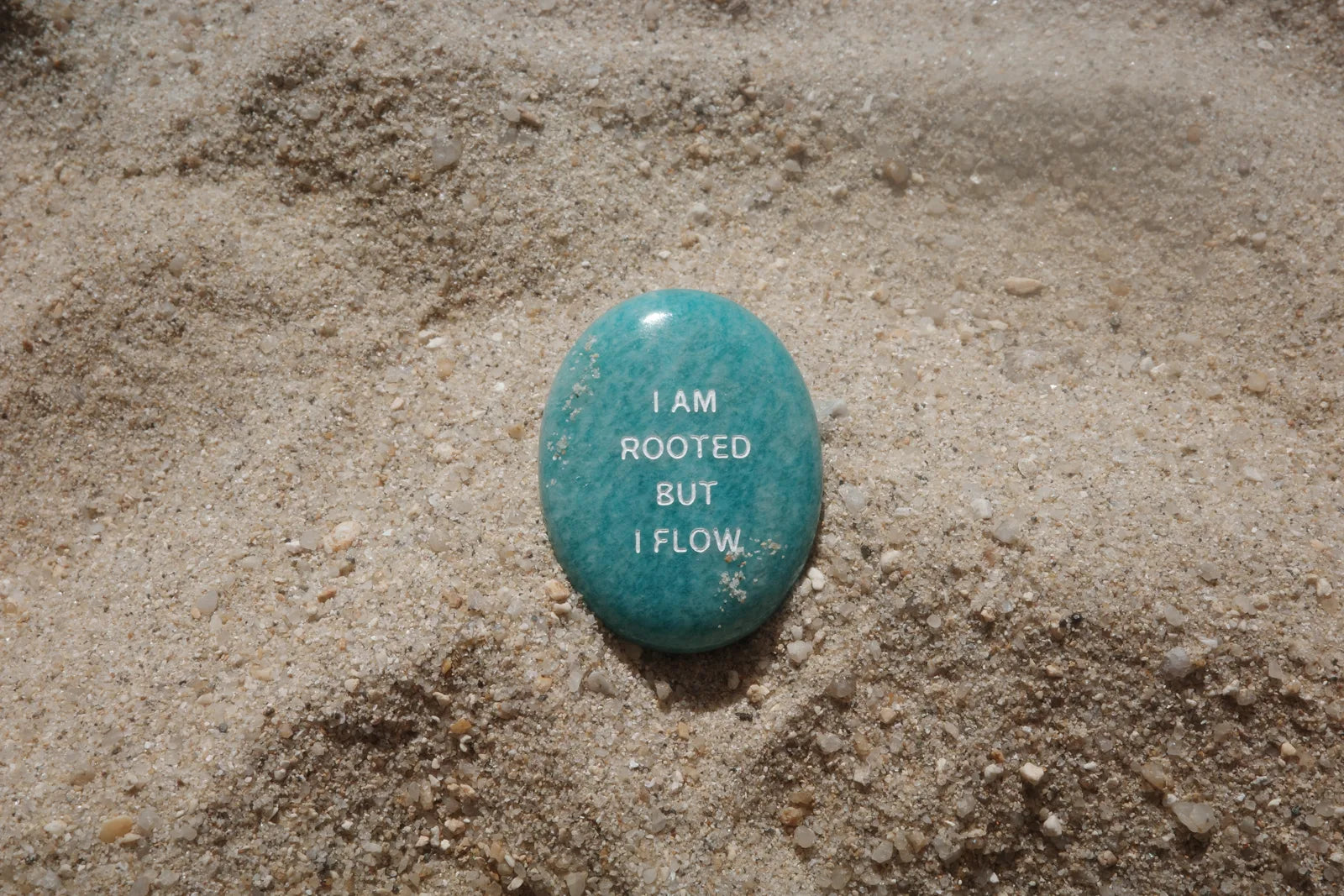An amazonite stone engraved with I Am Rooted But I Flow on beach sand